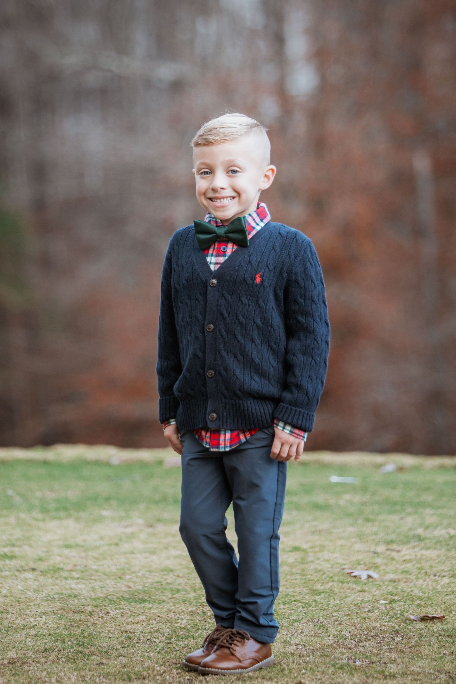 Young boy in navy cable-knit cardigan, plaid shirt, and green bow tie smiles outdoors on grass.