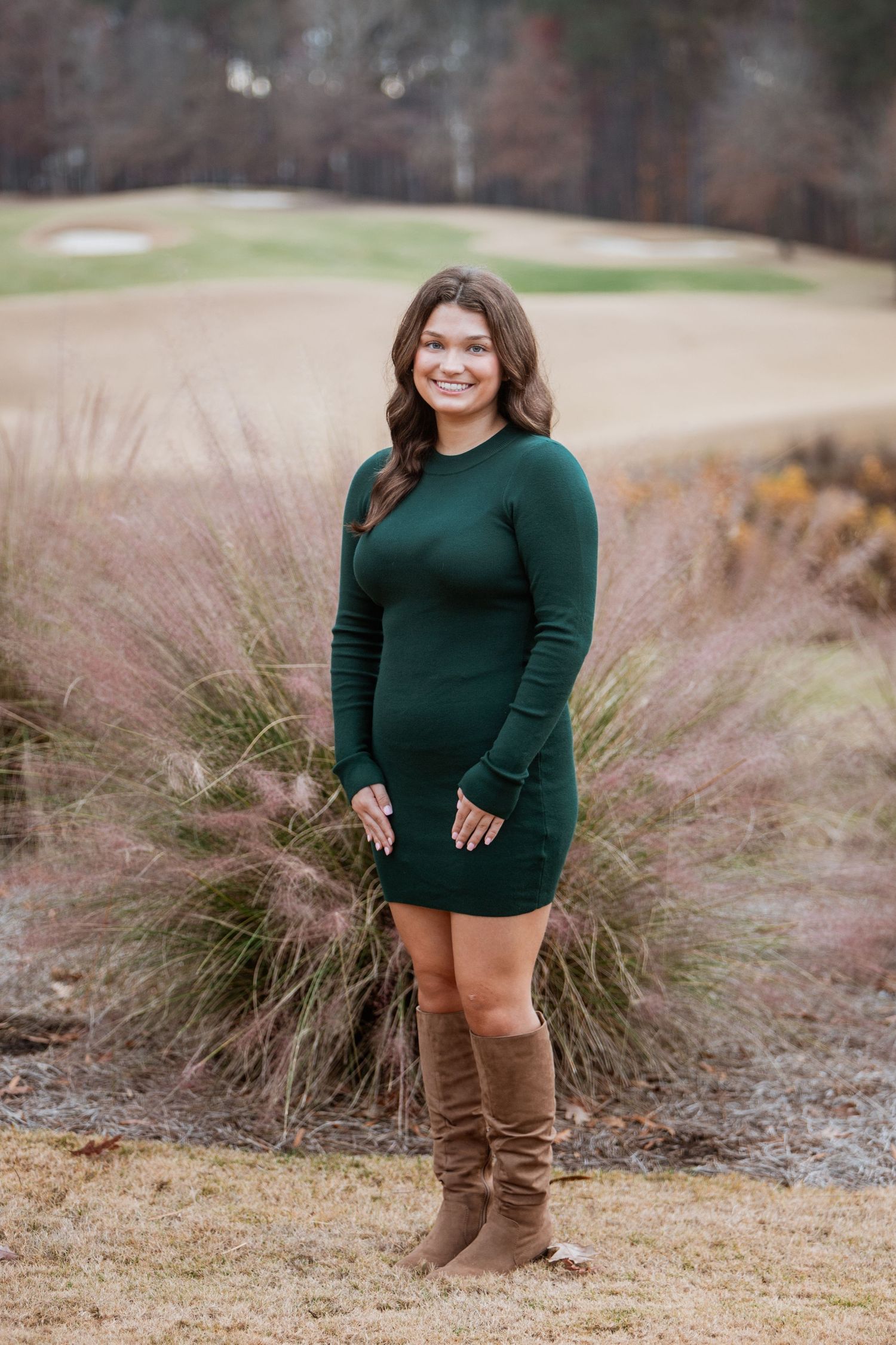 Young woman in green long-sleeve mini dress and brown boots on golf course.