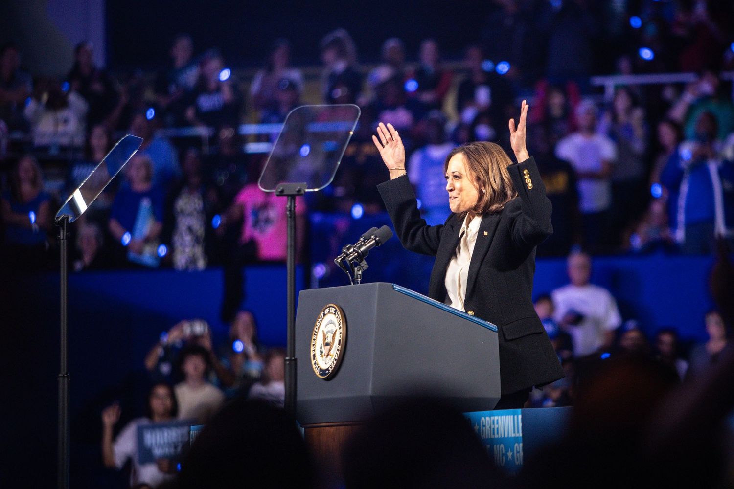 A woman in a black blazer raises both hands while speaking at a Harris-Walz presidential campaign rally.
