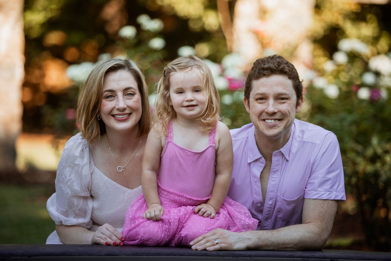 Smiling family of three posing outdoors with bokeh floral background, parents flanking happy toddler.