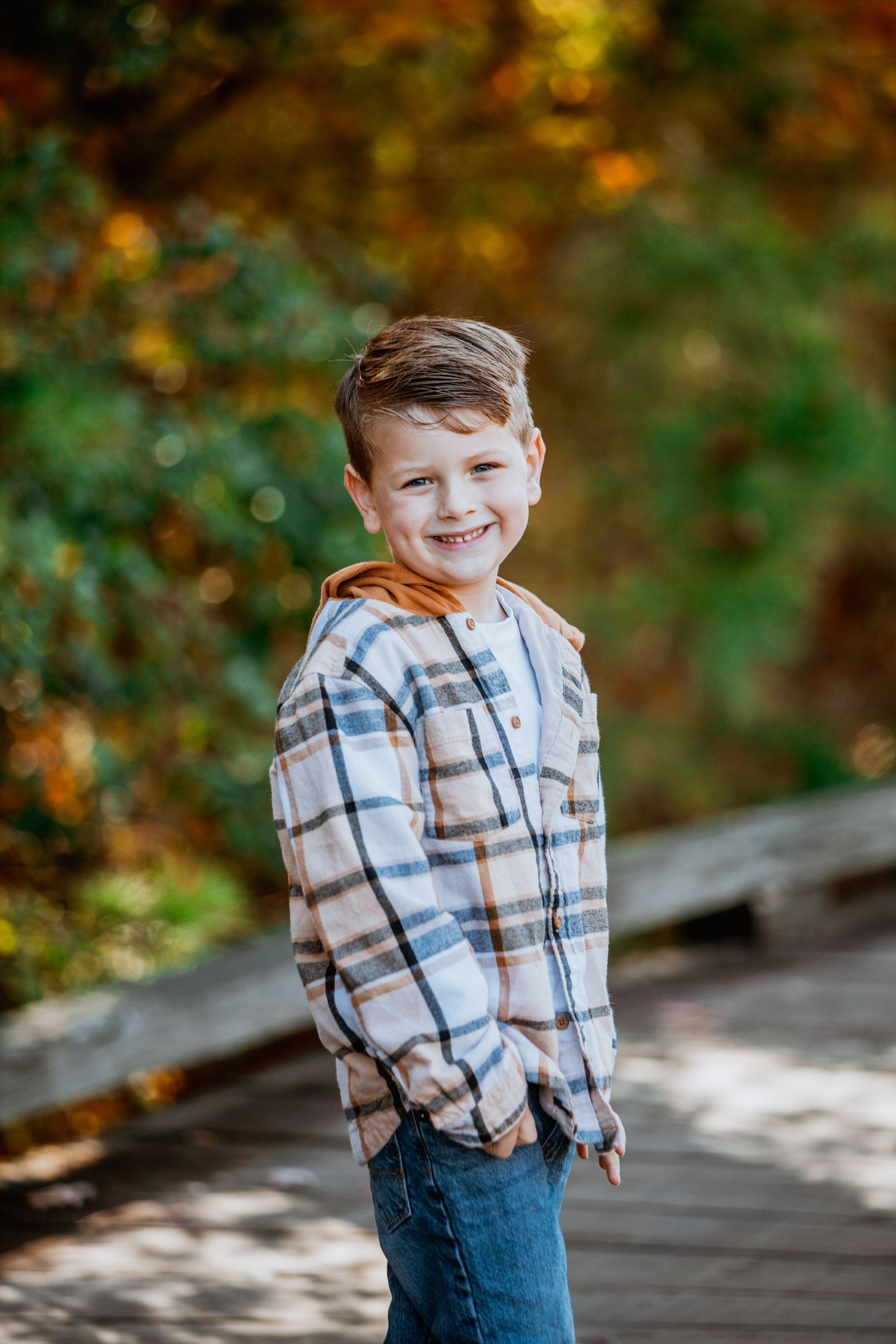 Smiling young boy in plaid flannel shirt and orange hoodie posing outdoors in autumn setting.