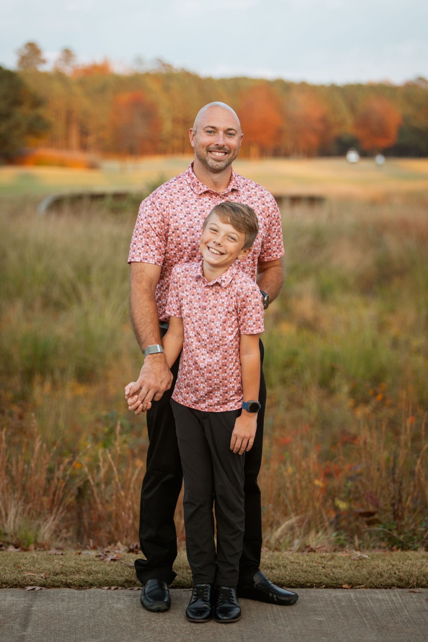Father and son wear matching pink floral shirts posing together in autumn outdoor portrait.