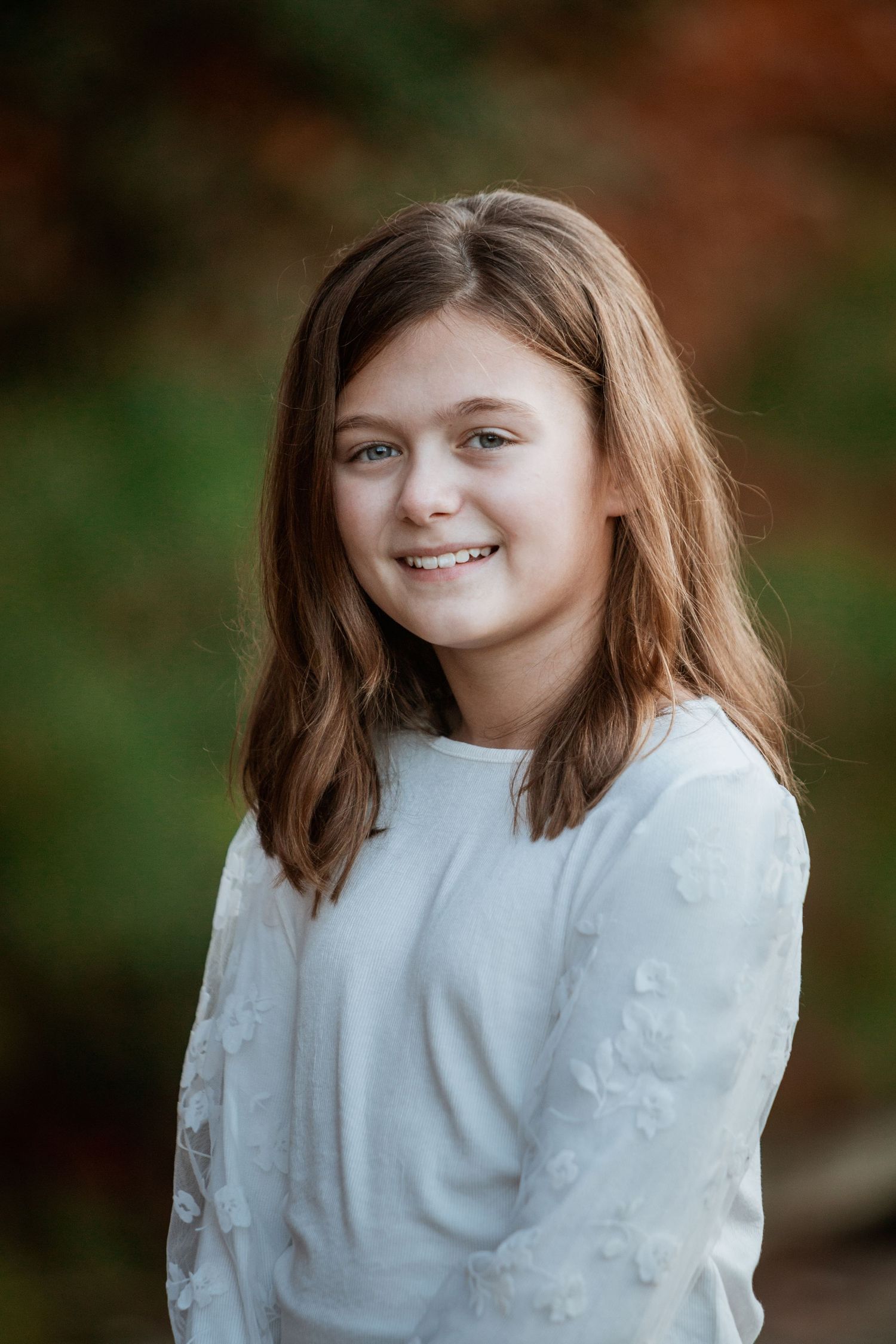 Smiling young girl with brown hair wearing white floral top in autumn outdoor portrait.