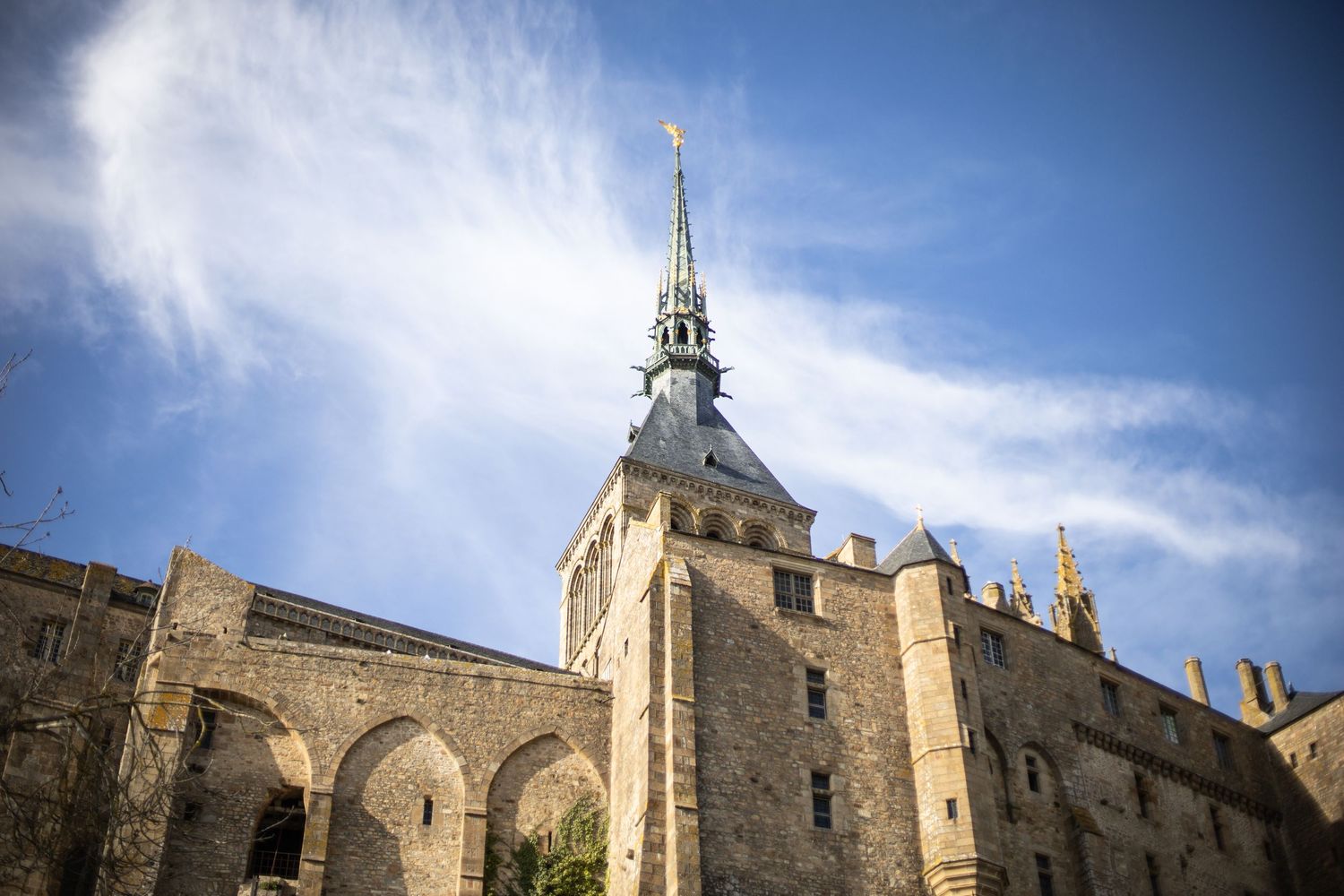 Low angle view of Mont Saint-Michel abbey tower and medieval stone walls against a blue sky.