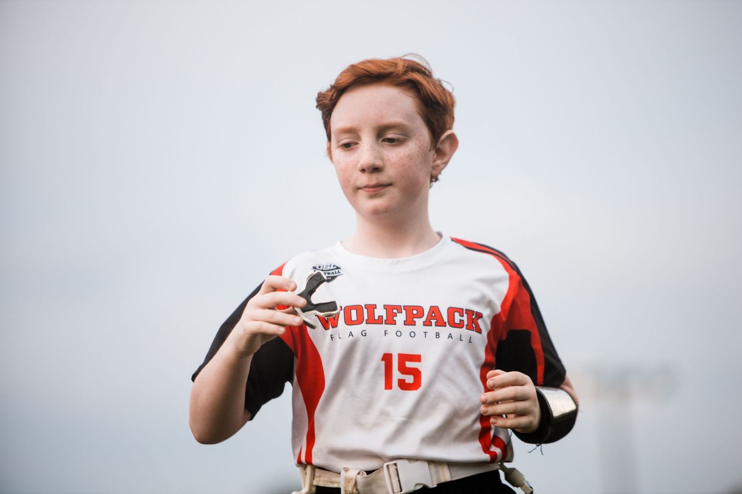 Young girl in Wolfpack #15 flag football jersey holding equipment outdoors.