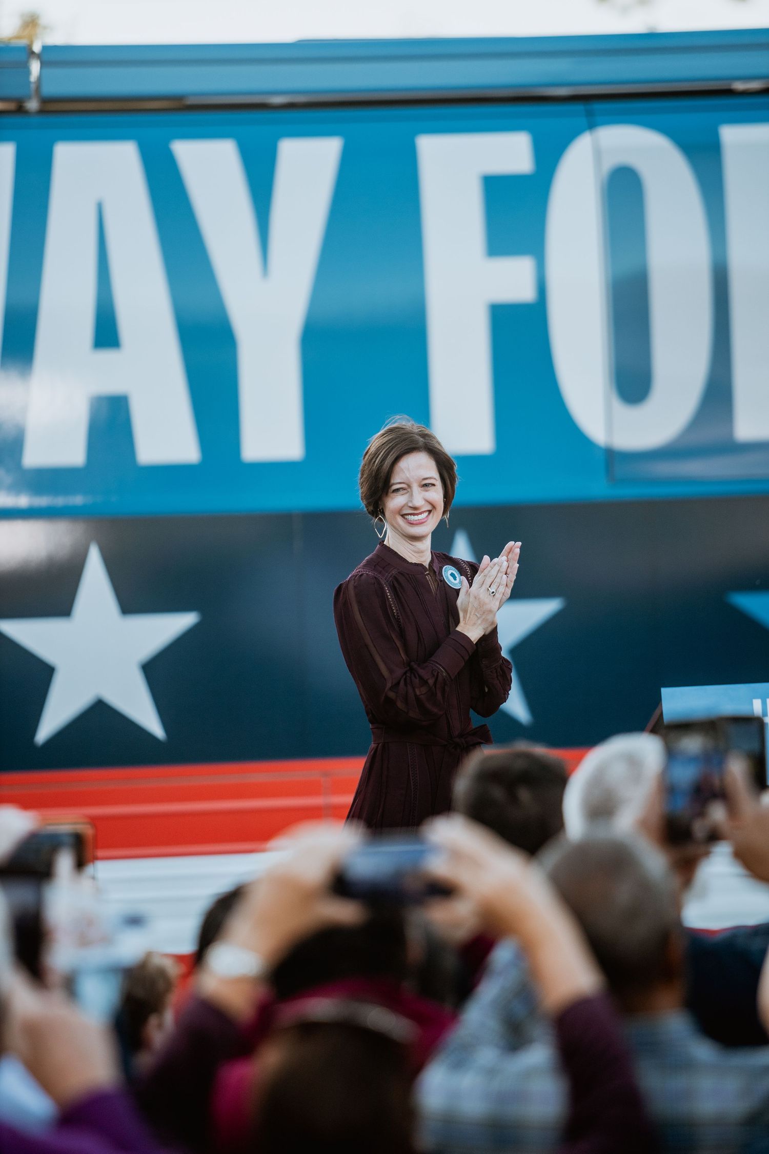 Woman in burgundy dress clapping at political campaign rally in front of a blue campaign bus with stars.