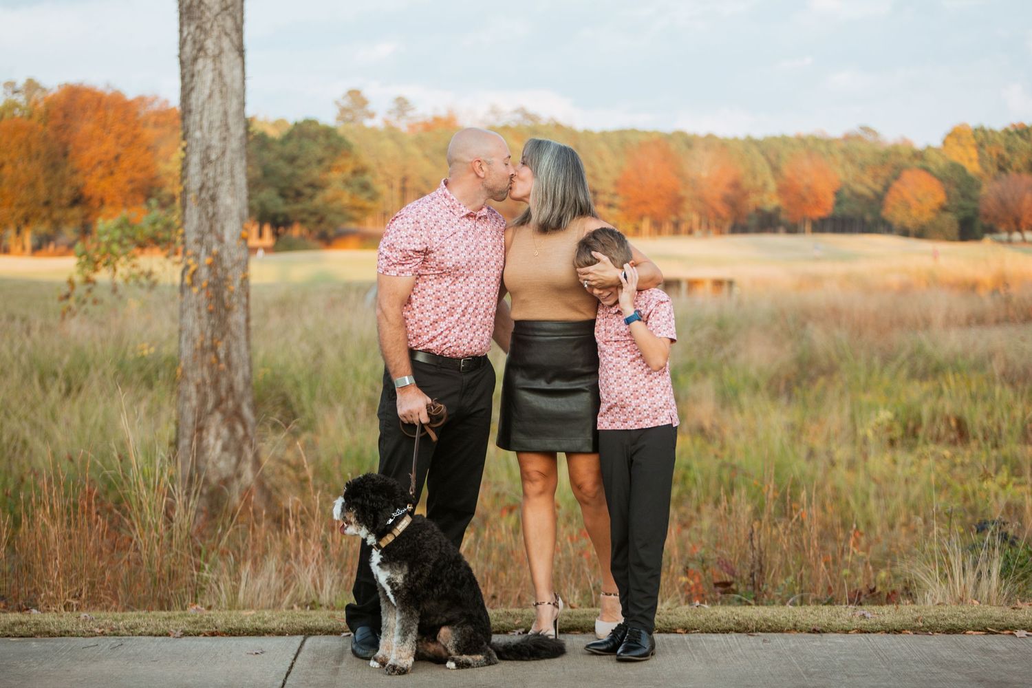 Family of three shares a kiss while son covers eyes with dog during fall photo.
