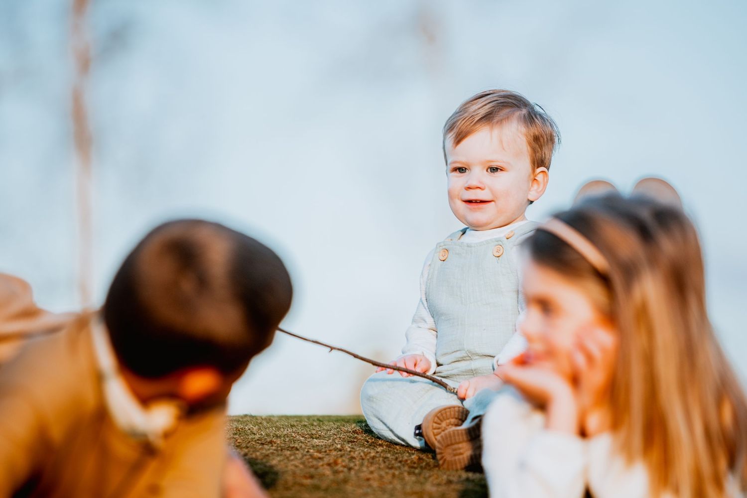 Adorable toddler sitting outdoors holding a stick, smiling joyfully with siblings in the foreground.