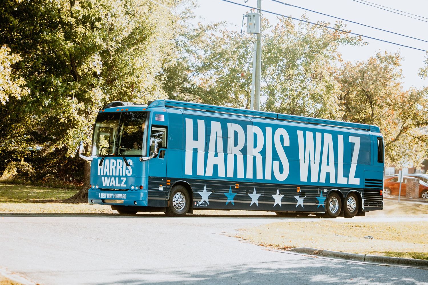 Blue Harris Walz 2024 presidential campaign bus parked on street near trees.