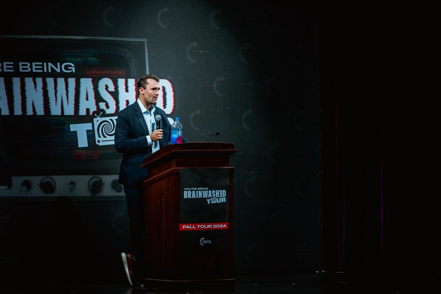Man in suit speaking at the Brainwashed Tour fall 2024 event at a podium.