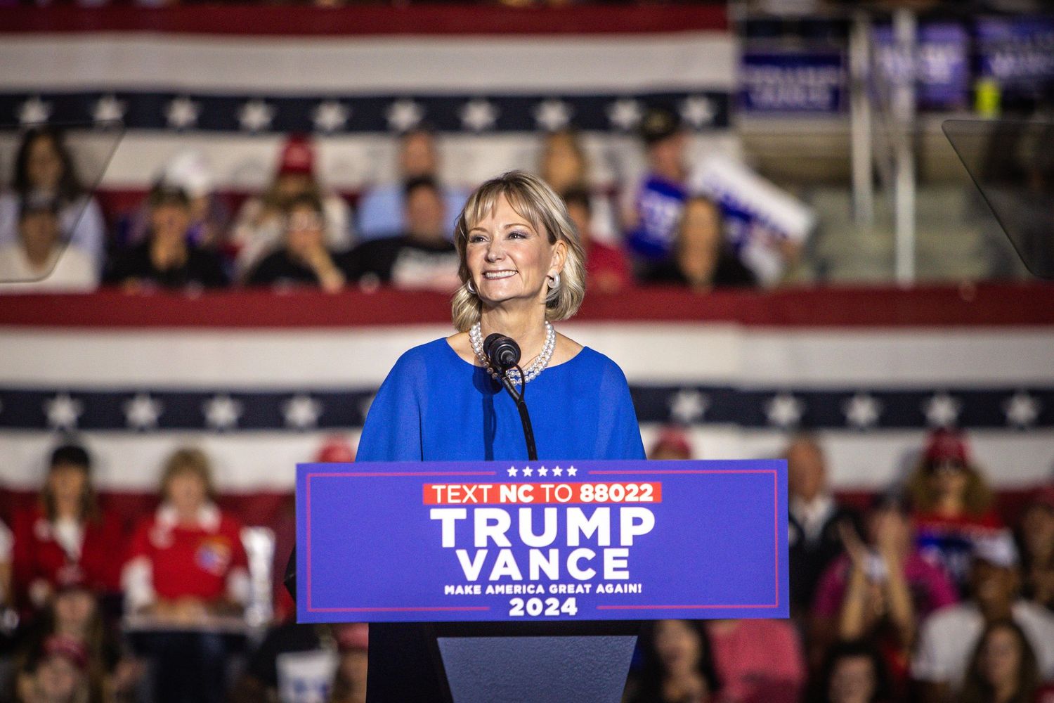 Woman in blue dress speaks at Trump Vance 2024 campaign rally before large crowd.