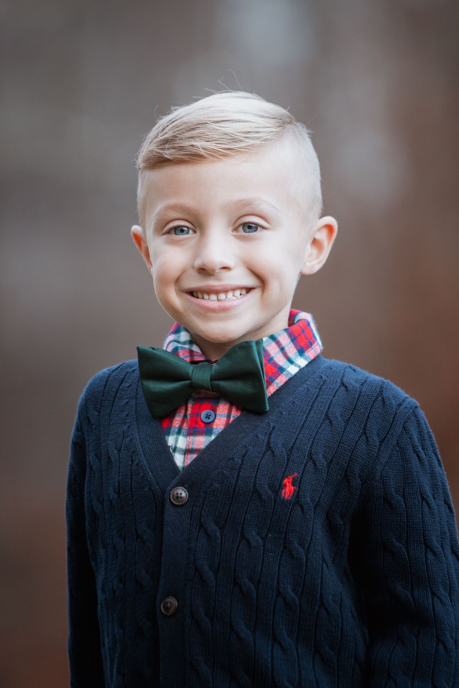 Close-up portrait of a smiling young boy in a navy cardigan, plaid shirt, and green bow tie.