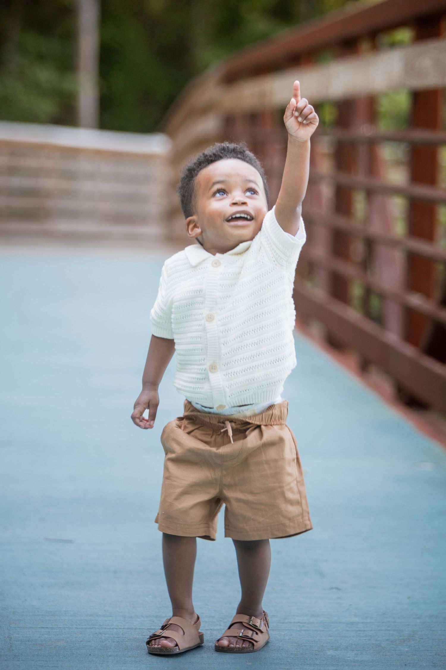 Excited toddler boy in white shirt and khaki shorts pointing upward while standing on a blue bridge.