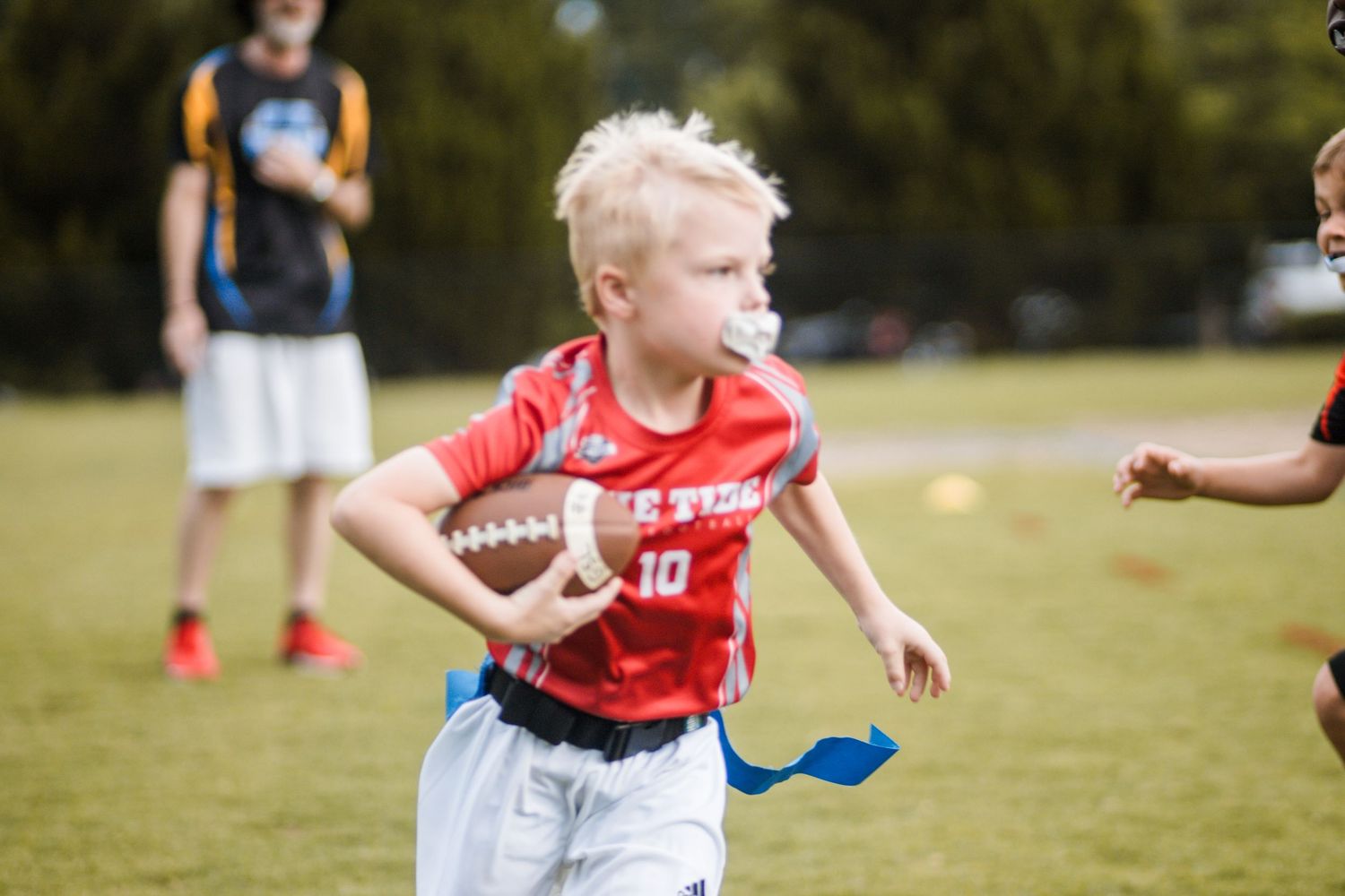 Blonde boy in red number 10 jersey runs with football during youth flag football game outdoors.
