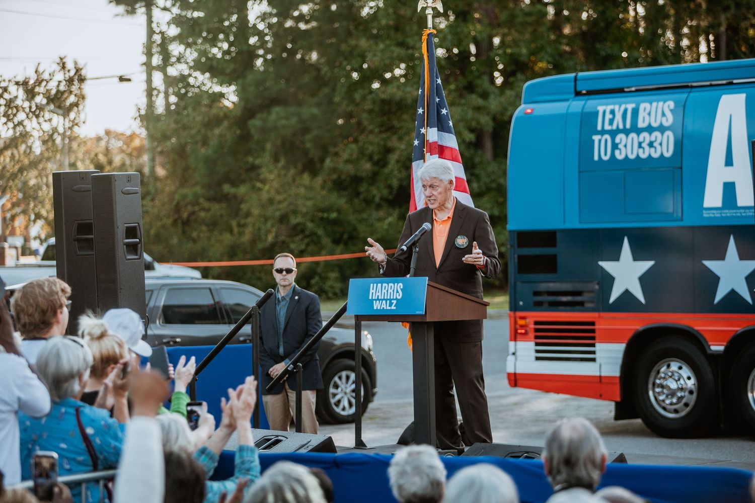 Speaker at Harris campaign rally podium with American flag and campaign bus reading TEXT BUS TO 30330 in background.