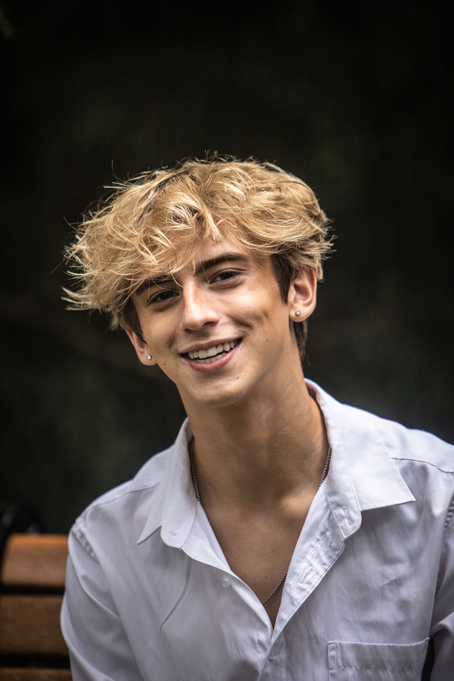 Smiling teenage boy with curly blonde hair wearing white shirt seated outdoors.