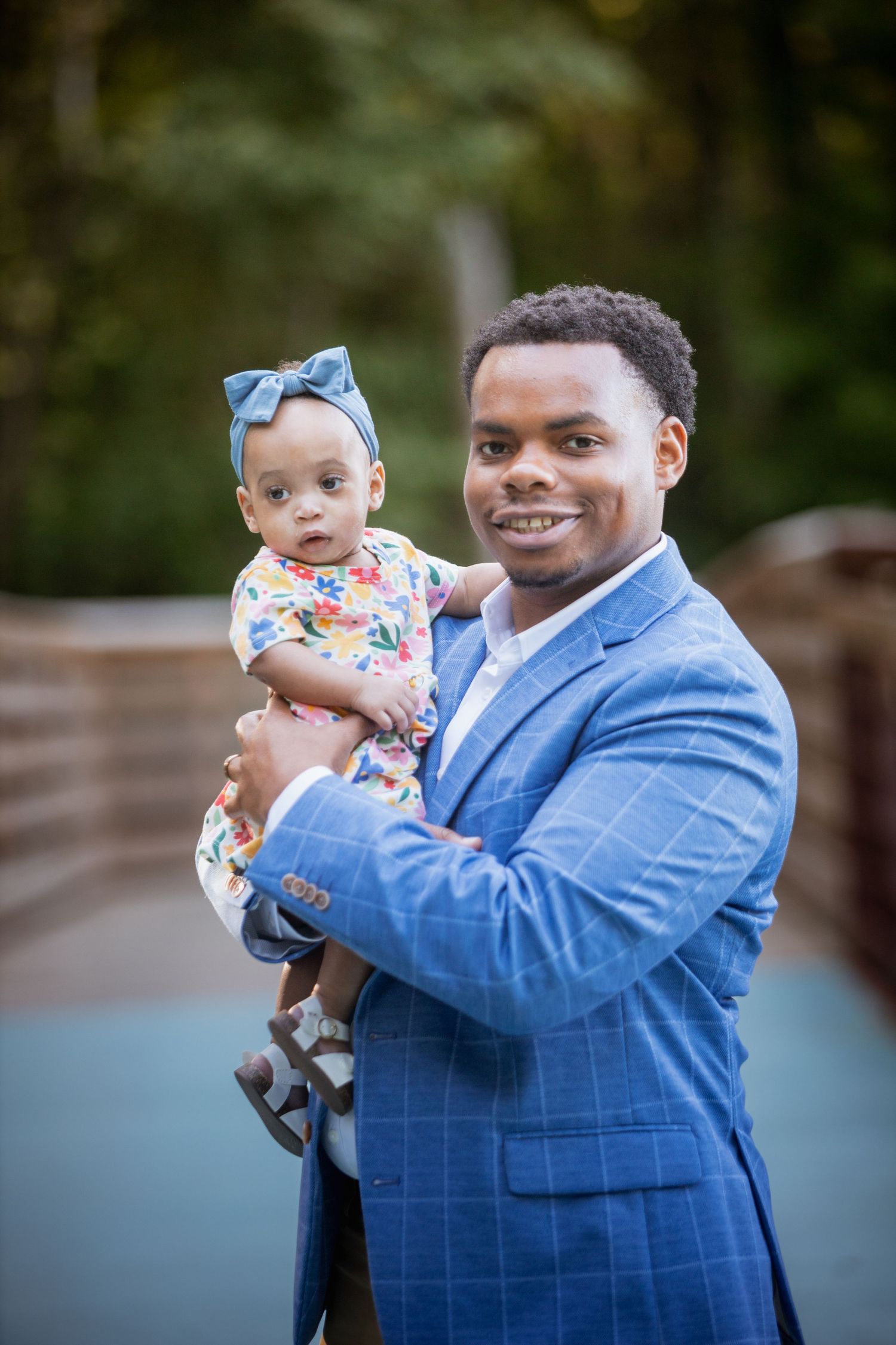 Proud father in blue plaid blazer holding his baby girl in colorful floral outfit on a bridge.