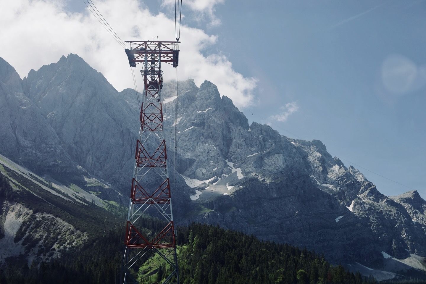 A red cable car pylon tower with jagged Alpine mountain peaks in the background.