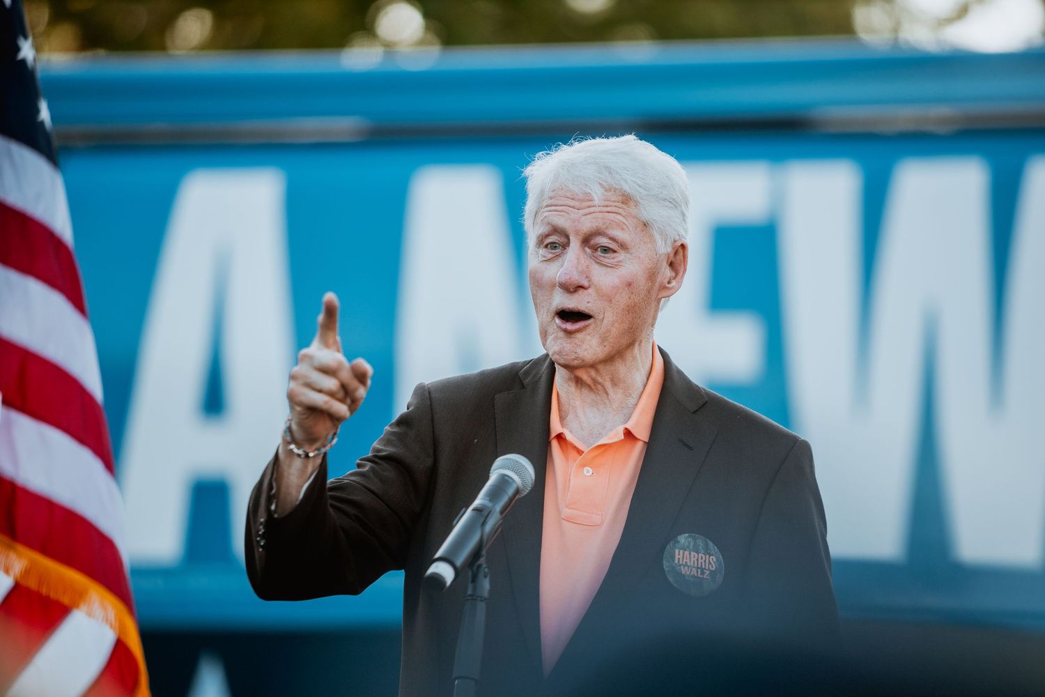 Senior man in dark blazer points finger while speaking at outdoor Harris campaign rally with blue banner.