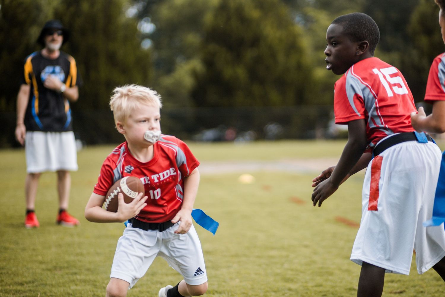 Blonde boy in red jersey carries football past defender in youth flag football game on grass field.