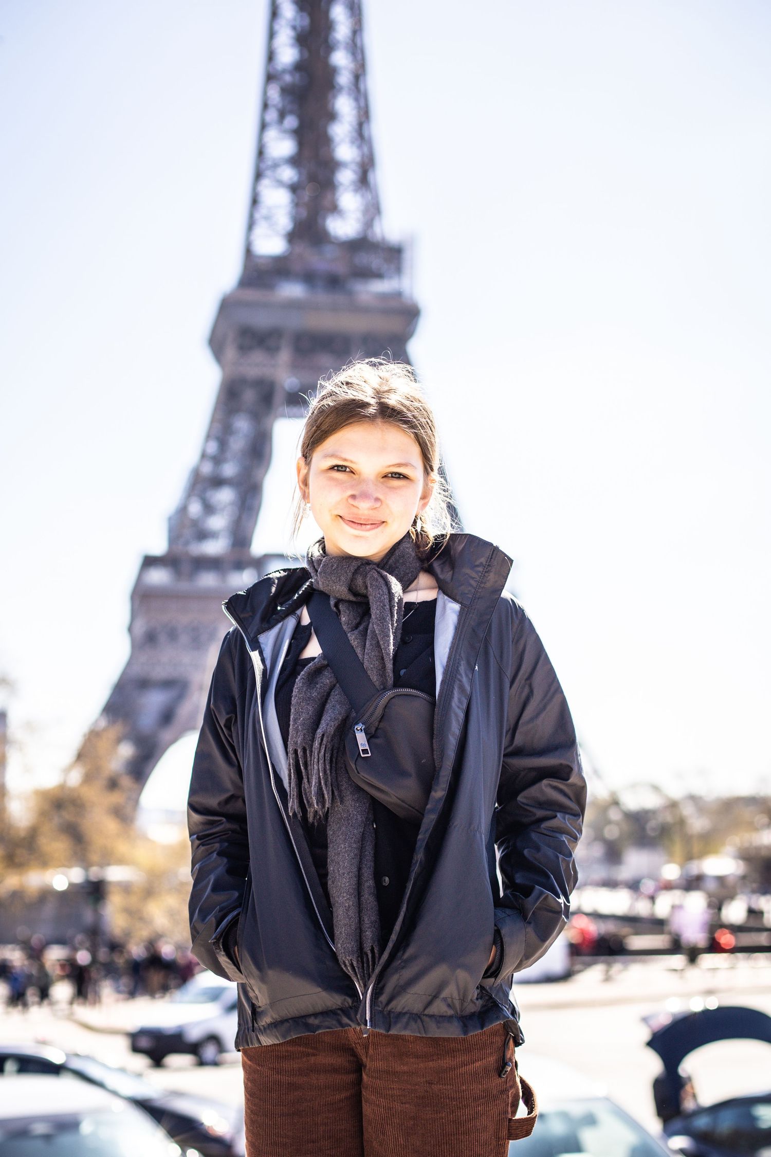 Smiling young woman in a dark jacket standing confidently in front of the Eiffel Tower in Paris.