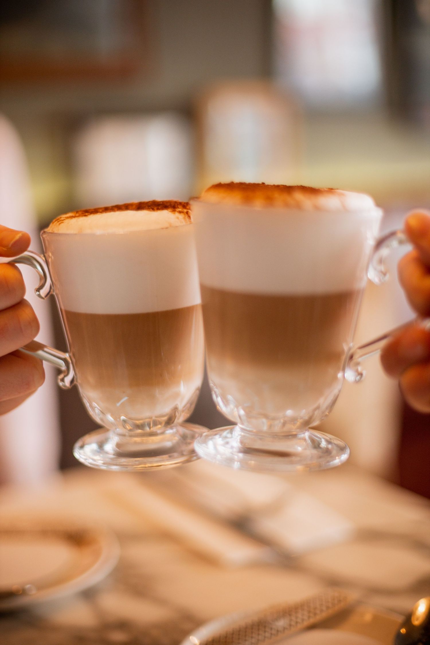 Two hands holding tall glass mugs of layered latte macchiato coffee with frothy milk foam tops.