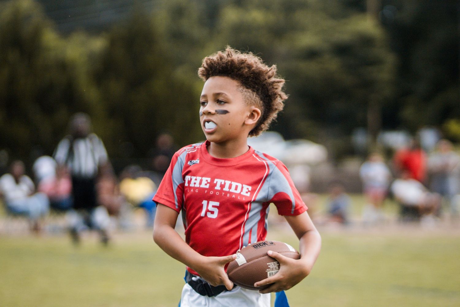 Young boy number 15 in red The Tide jersey smiling while holding football on field.