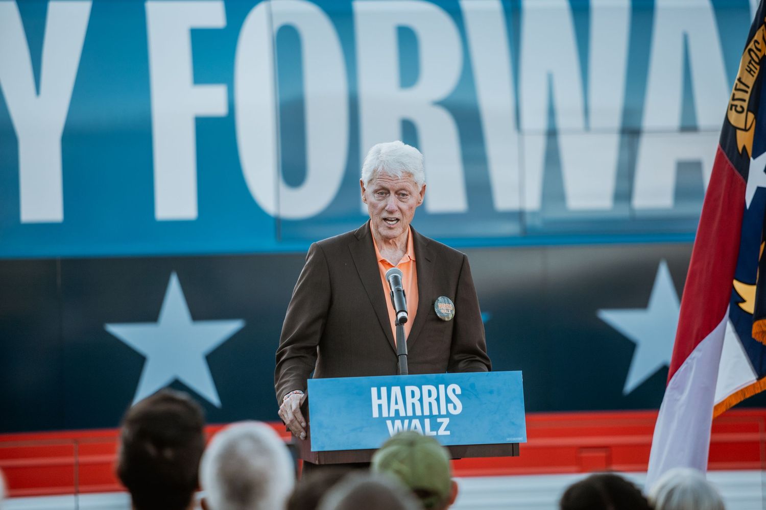 Senior man in dark blazer speaks passionately at Harris-Walz campaign podium with Forward banner behind.