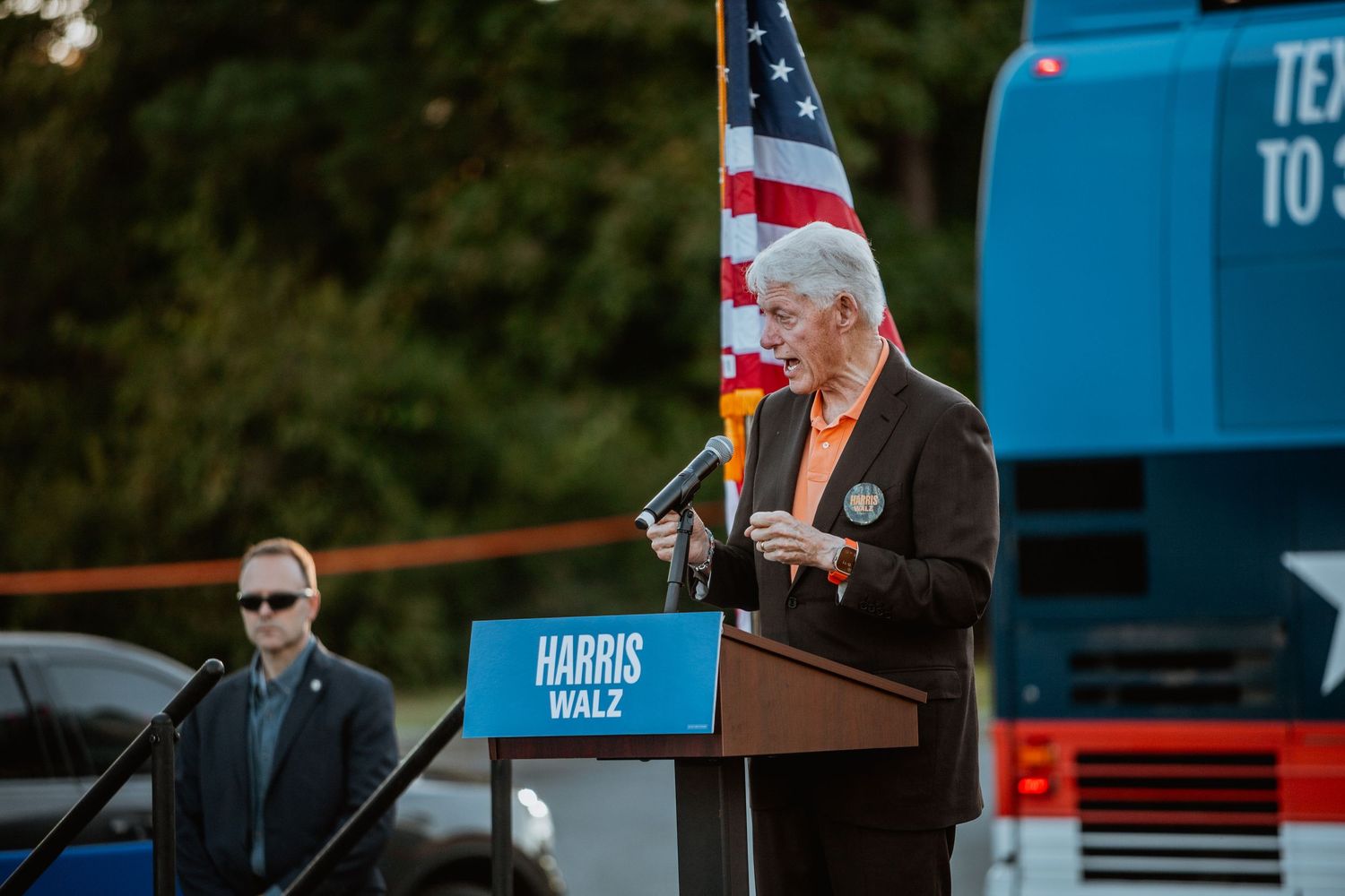 Elderly speaker at Harris Walz campaign podium with American flag outdoors.