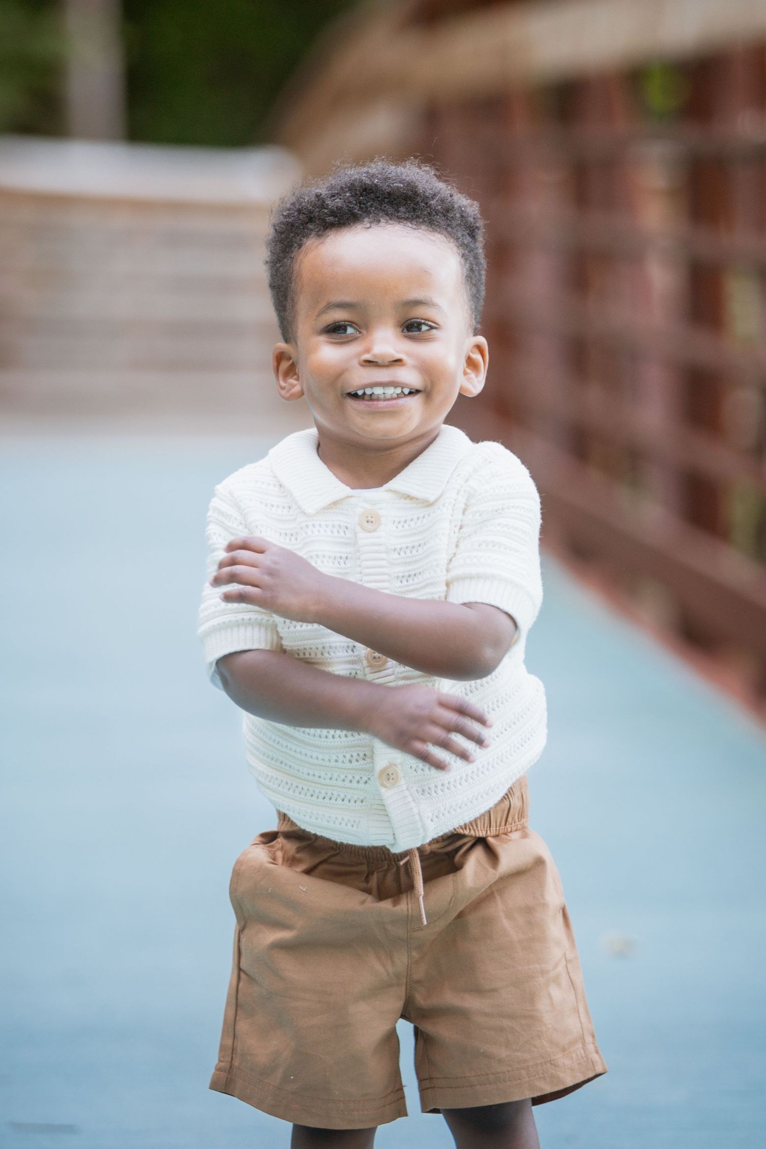 Happy young boy in white knit top and brown shorts posing with arms crossed on an outdoor bridge.