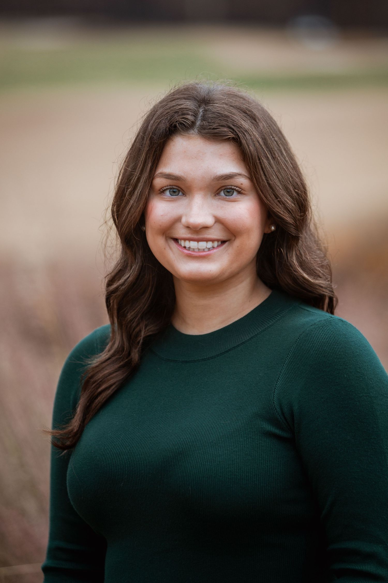 Young woman with brown hair smiling outdoors in a green sweater during a fall portrait session.