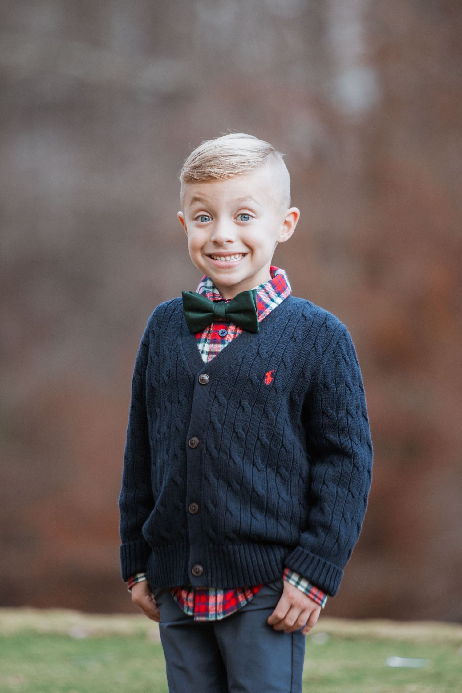 Smiling young boy in navy cardigan, plaid shirt, and green bow tie poses against a brick wall.