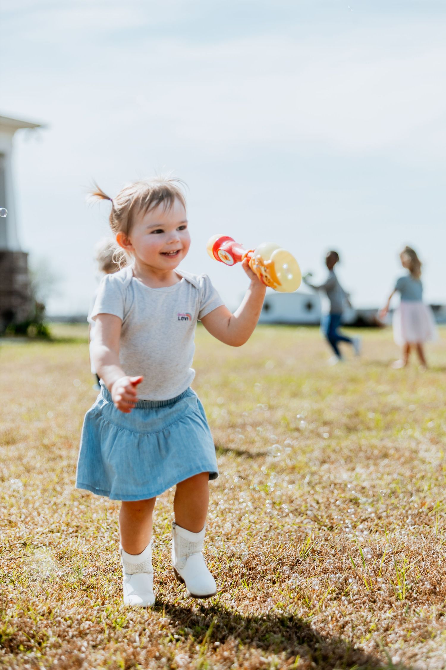 Happy toddler girl blowing bubbles in a sunny backyard while wearing a denim skirt.