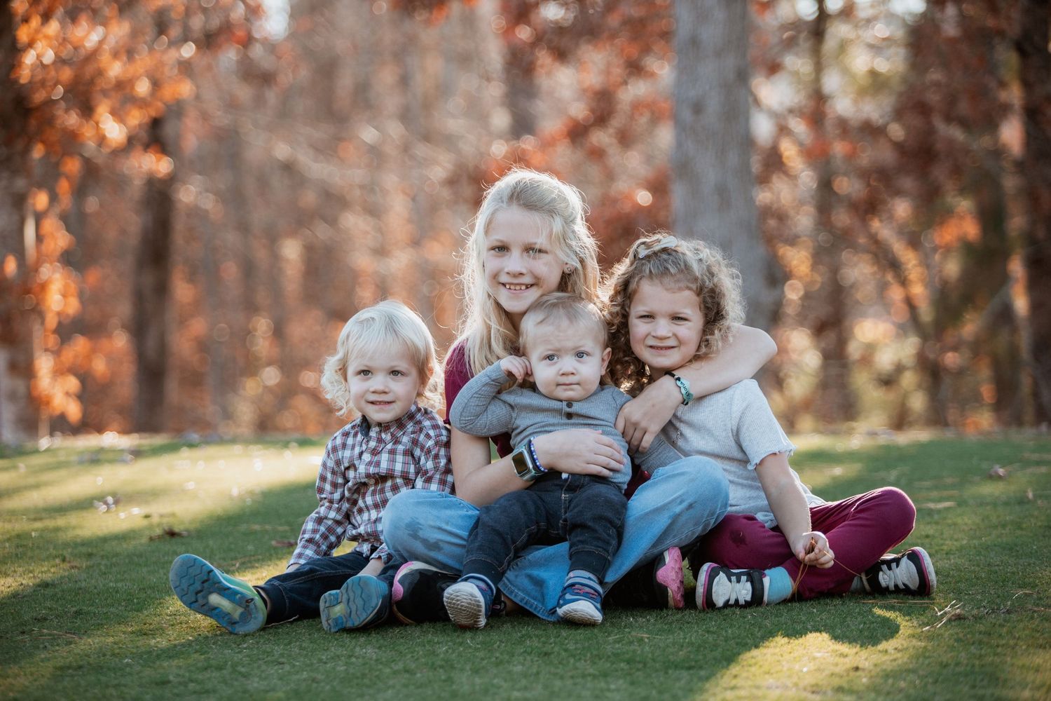 Four young siblings sit closely together on green grass with colorful autumn trees behind them.