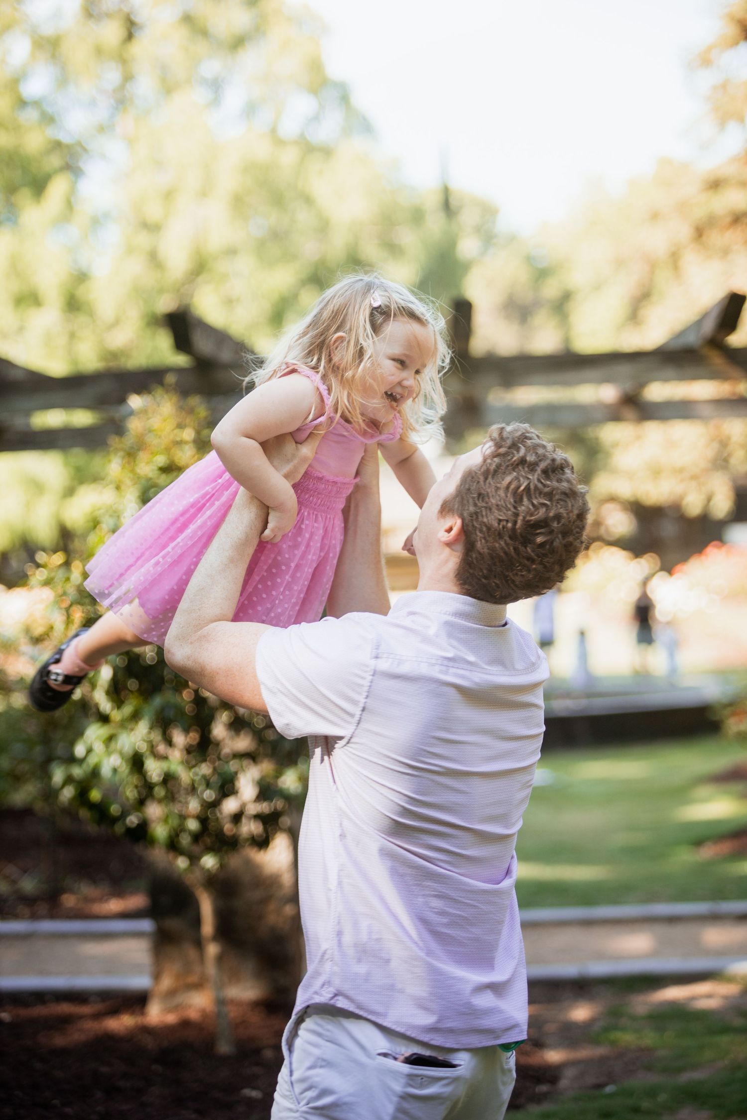 Father lifting laughing young daughter in pink dress high in the air outdoors.