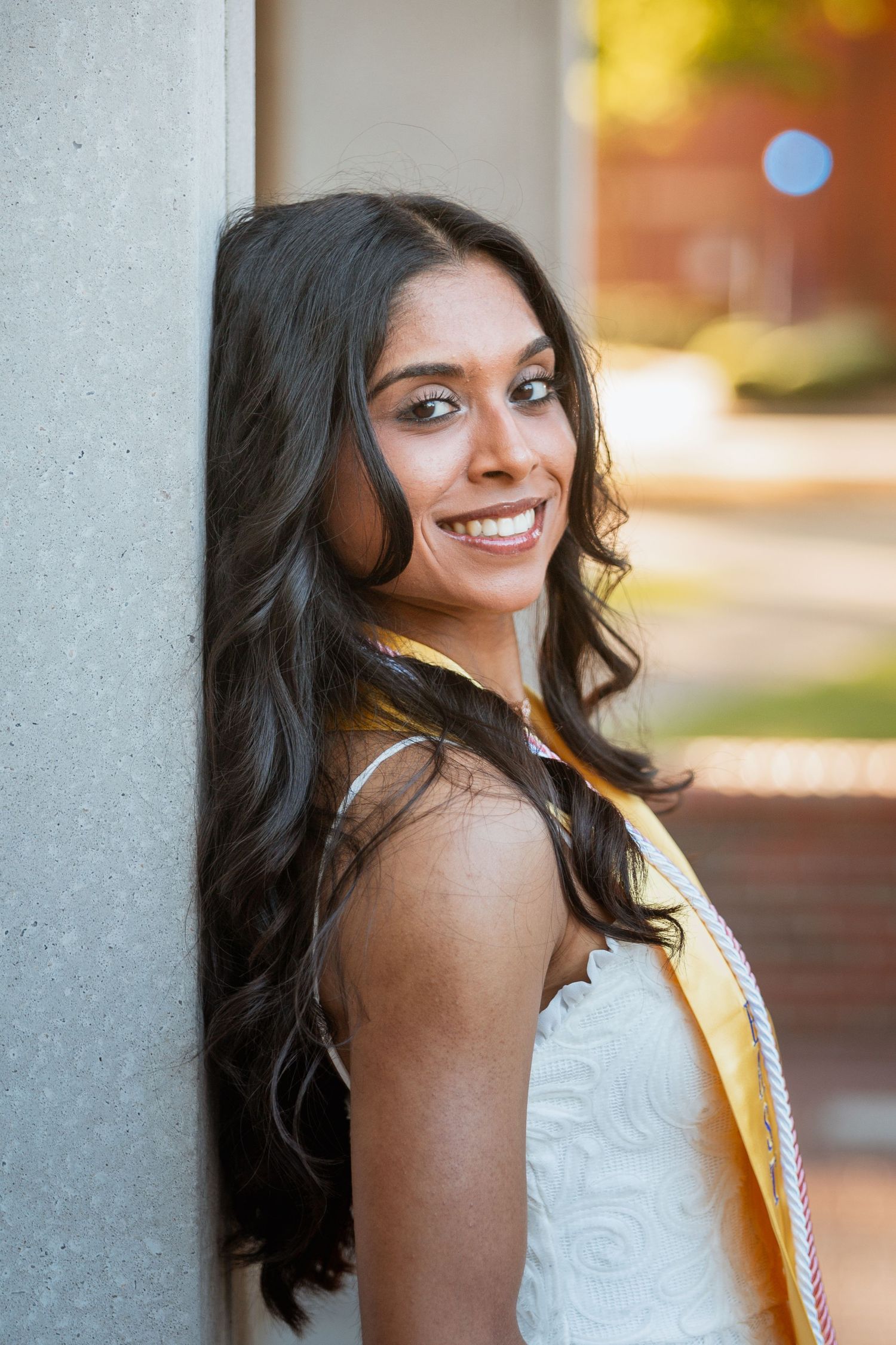 Smiling young woman in white dress and gold graduation stole leans against a concrete pillar.