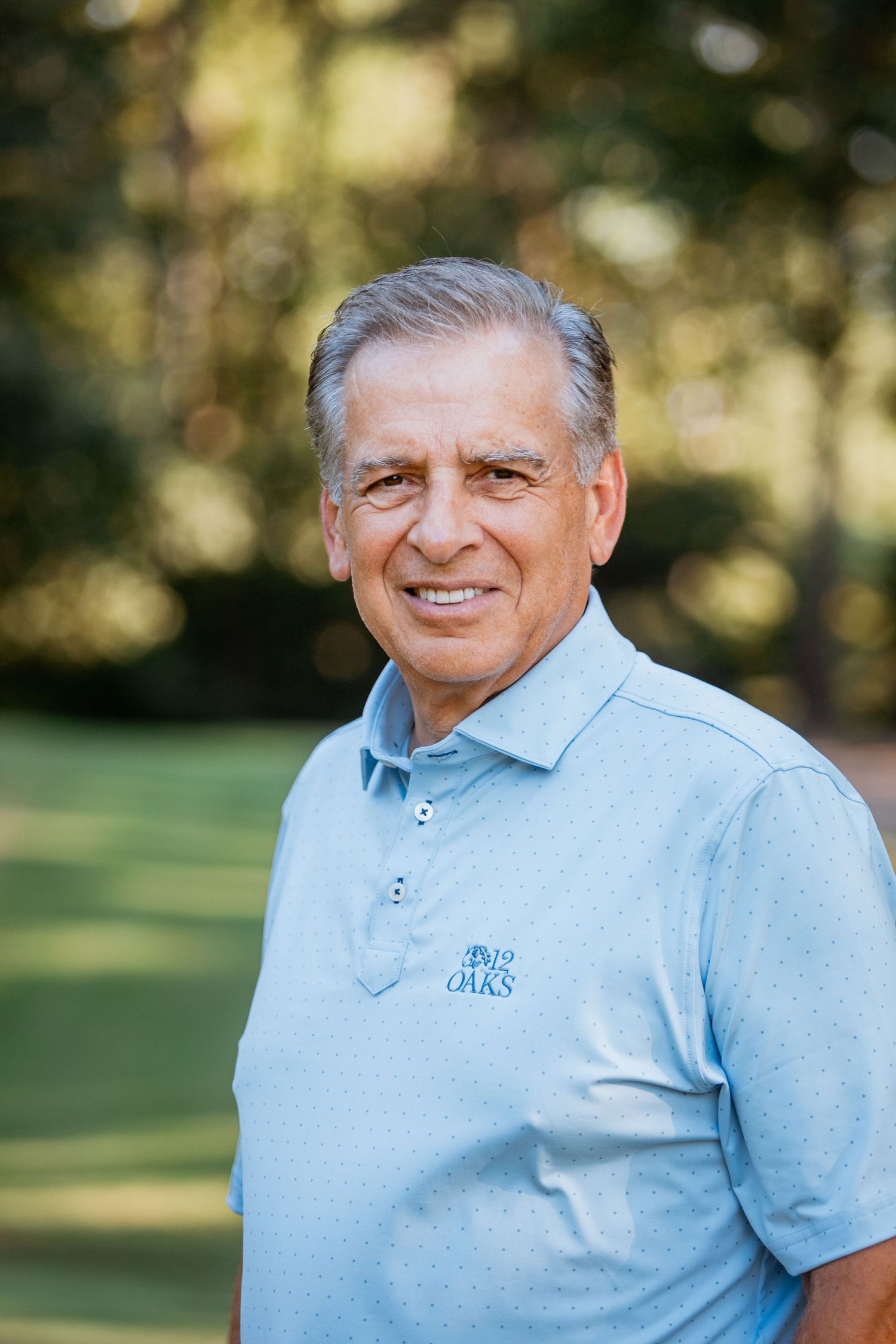Older man smiling outdoors wearing light blue golf polo shirt near trees.