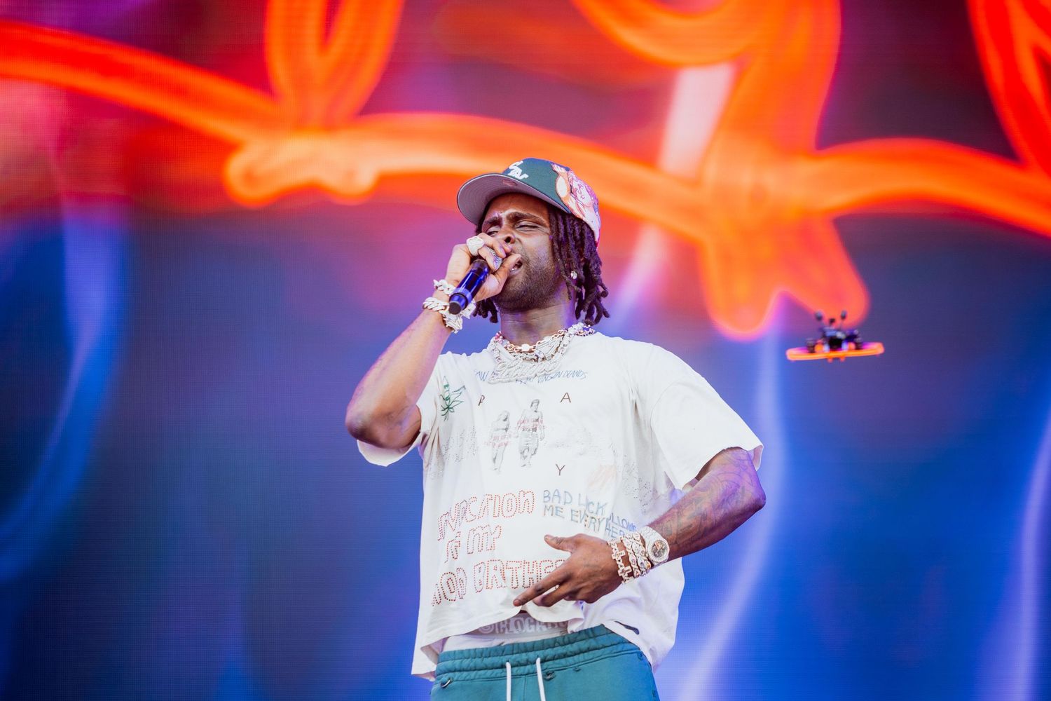 Male hip-hop artist performing on stage in a white tee and teal pants against a colorful neon backdrop.