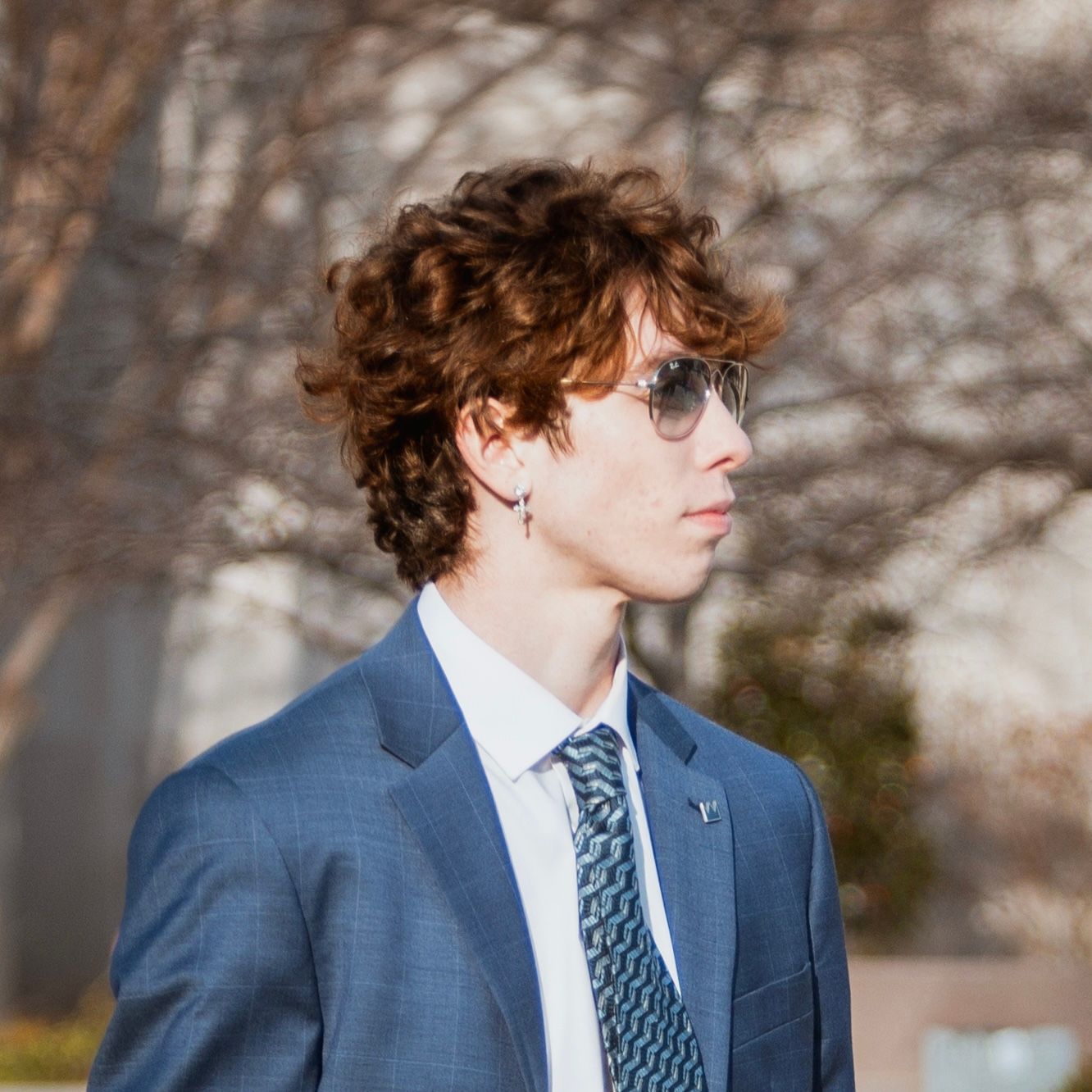 Stylish young man with curly hair in blue suit and aviator sunglasses posing outdoors.