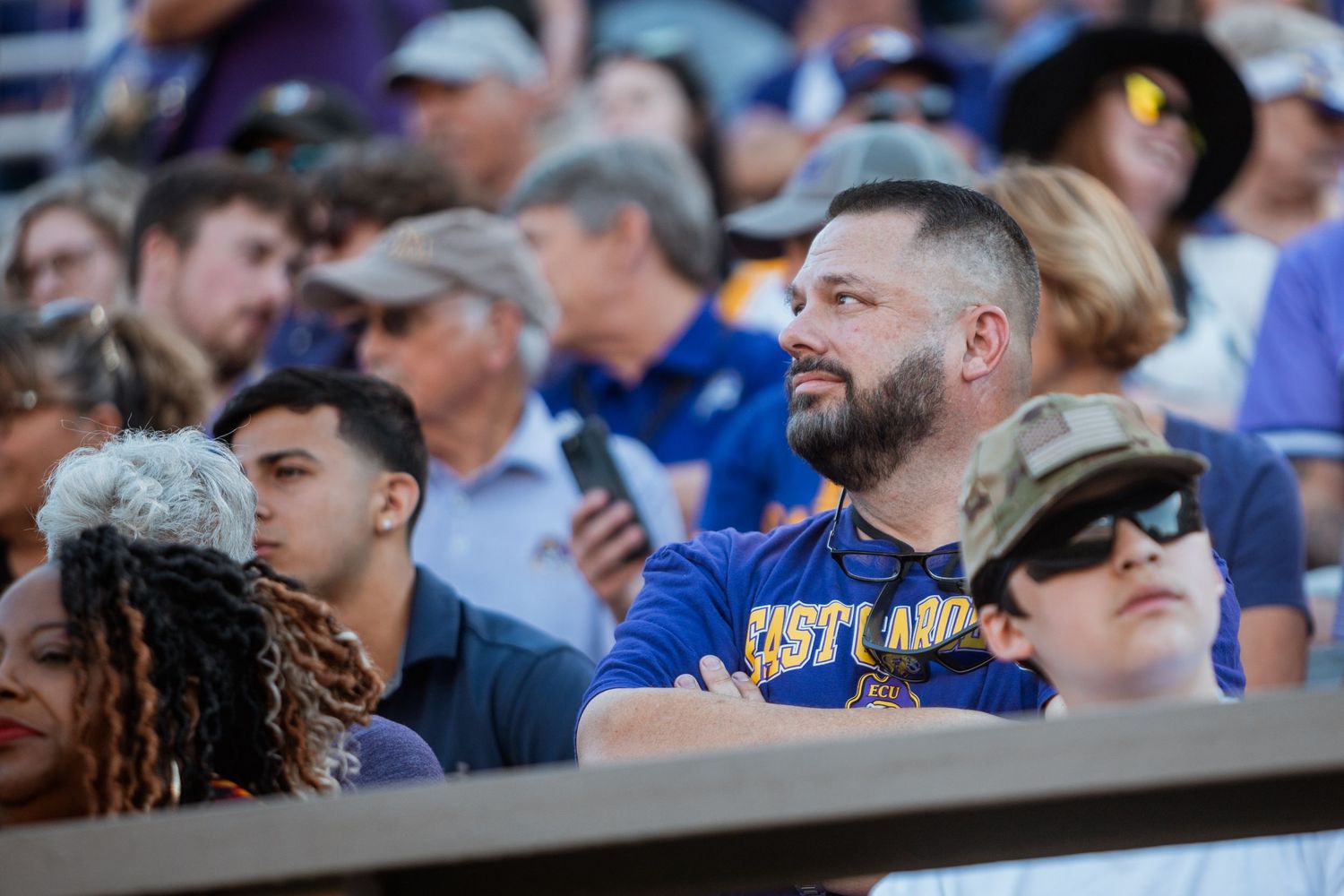 A fan wearing an ECU Pirates shirt watches the football game from the stands.