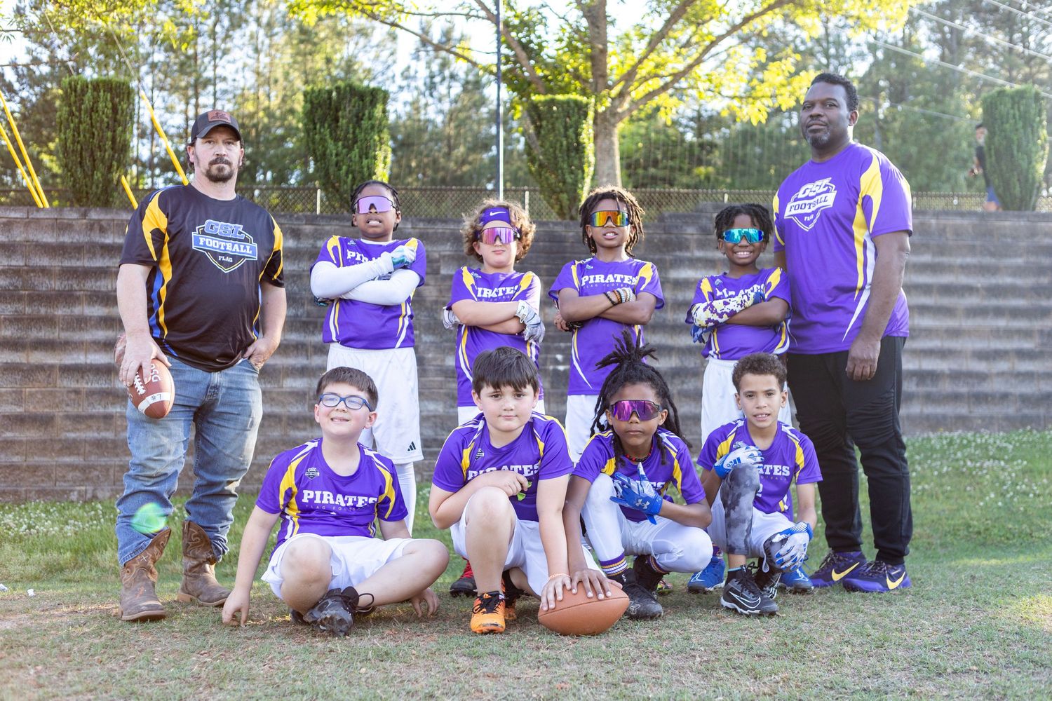 Youth flag football team in purple Pirates jerseys poses with coaches for team photo outdoors.