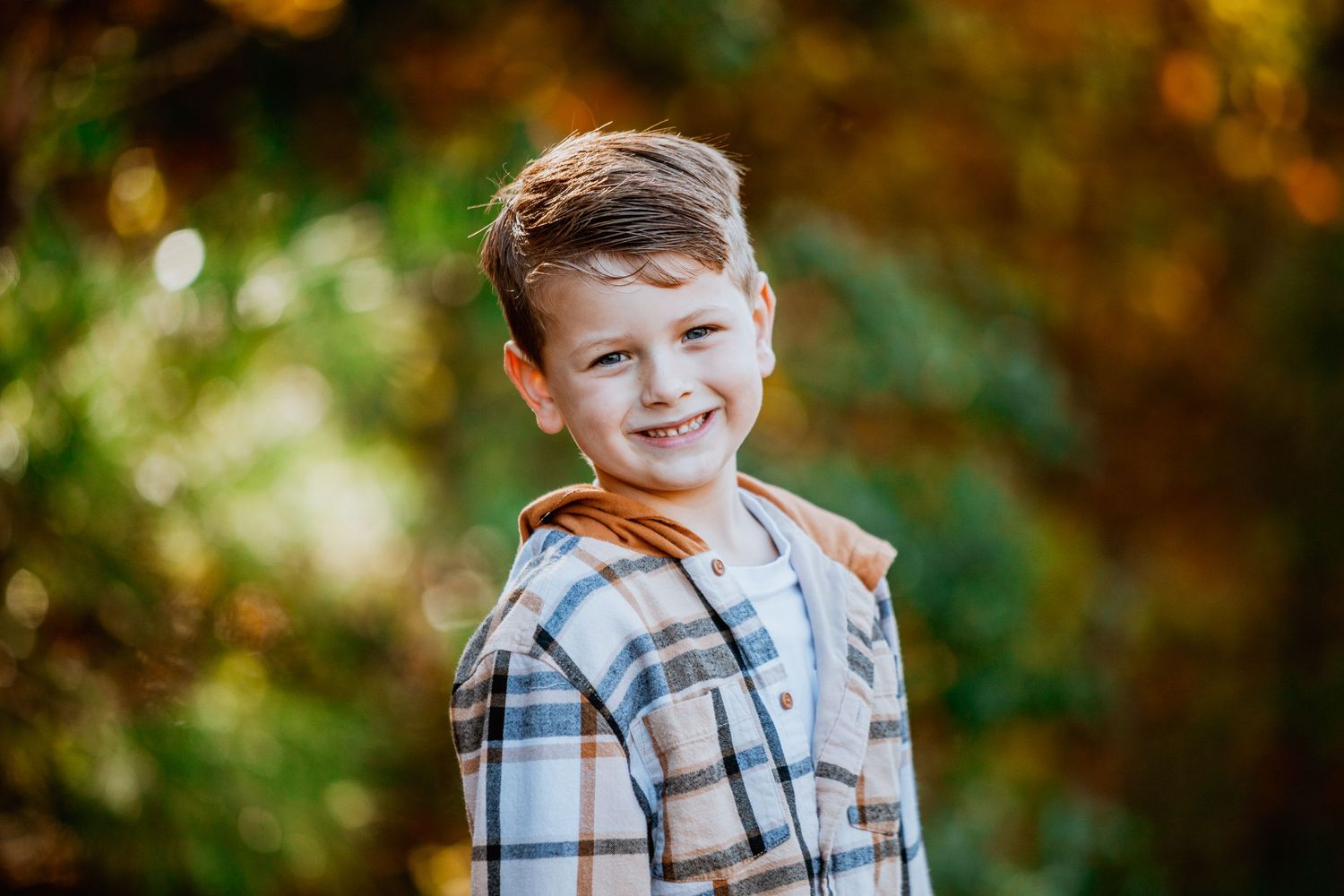 Smiling young boy in a plaid flannel shirt posing outdoors with colorful autumn foliage in the background.