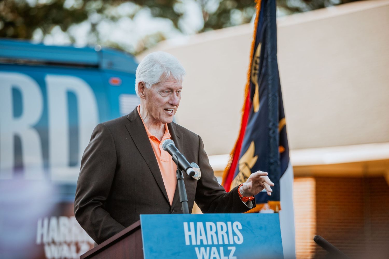 Older man in orange shirt and dark blazer speaks at Harris-Walz campaign podium at outdoor rally.