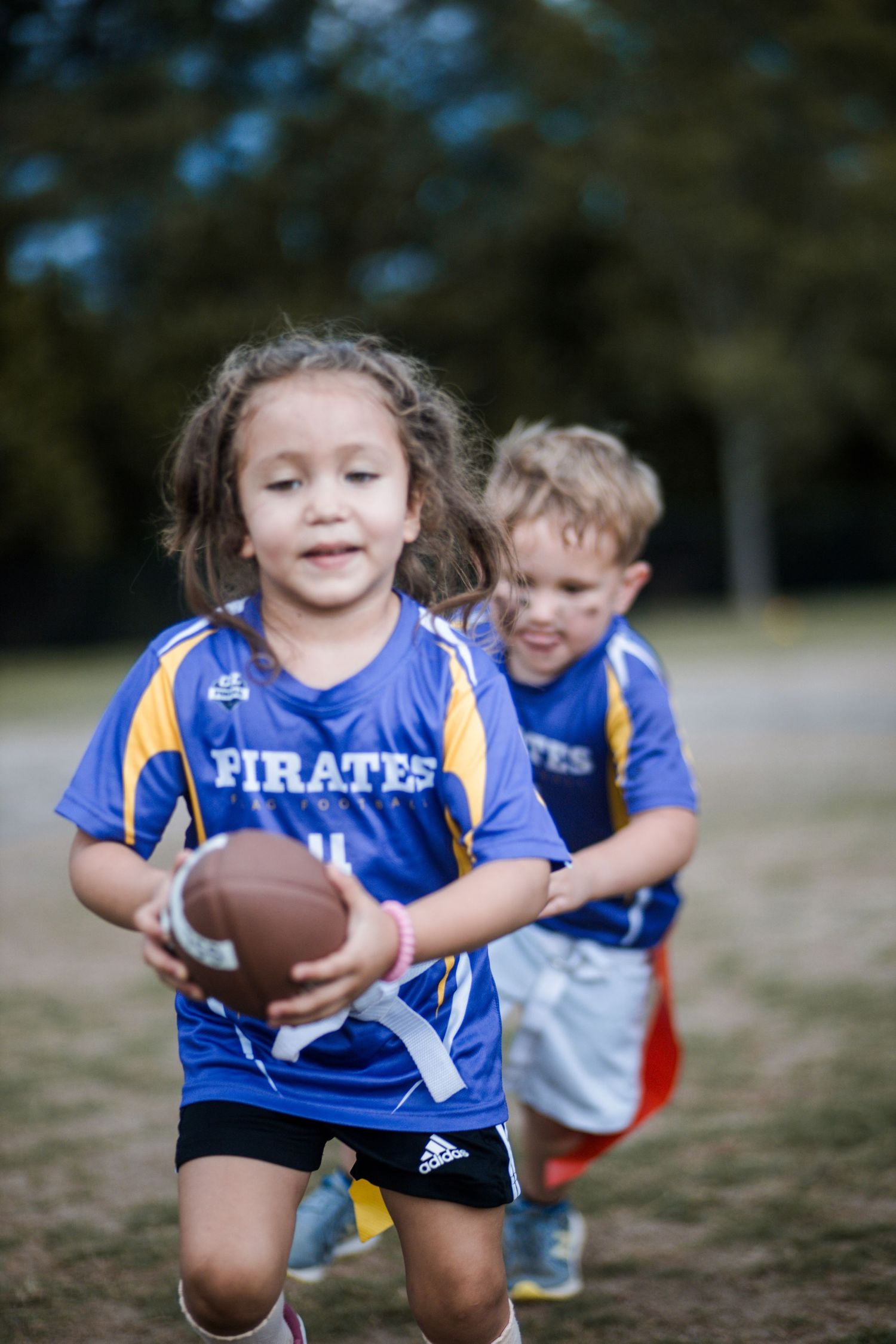 Young girl in blue Pirates jersey carrying football during youth flag football game.