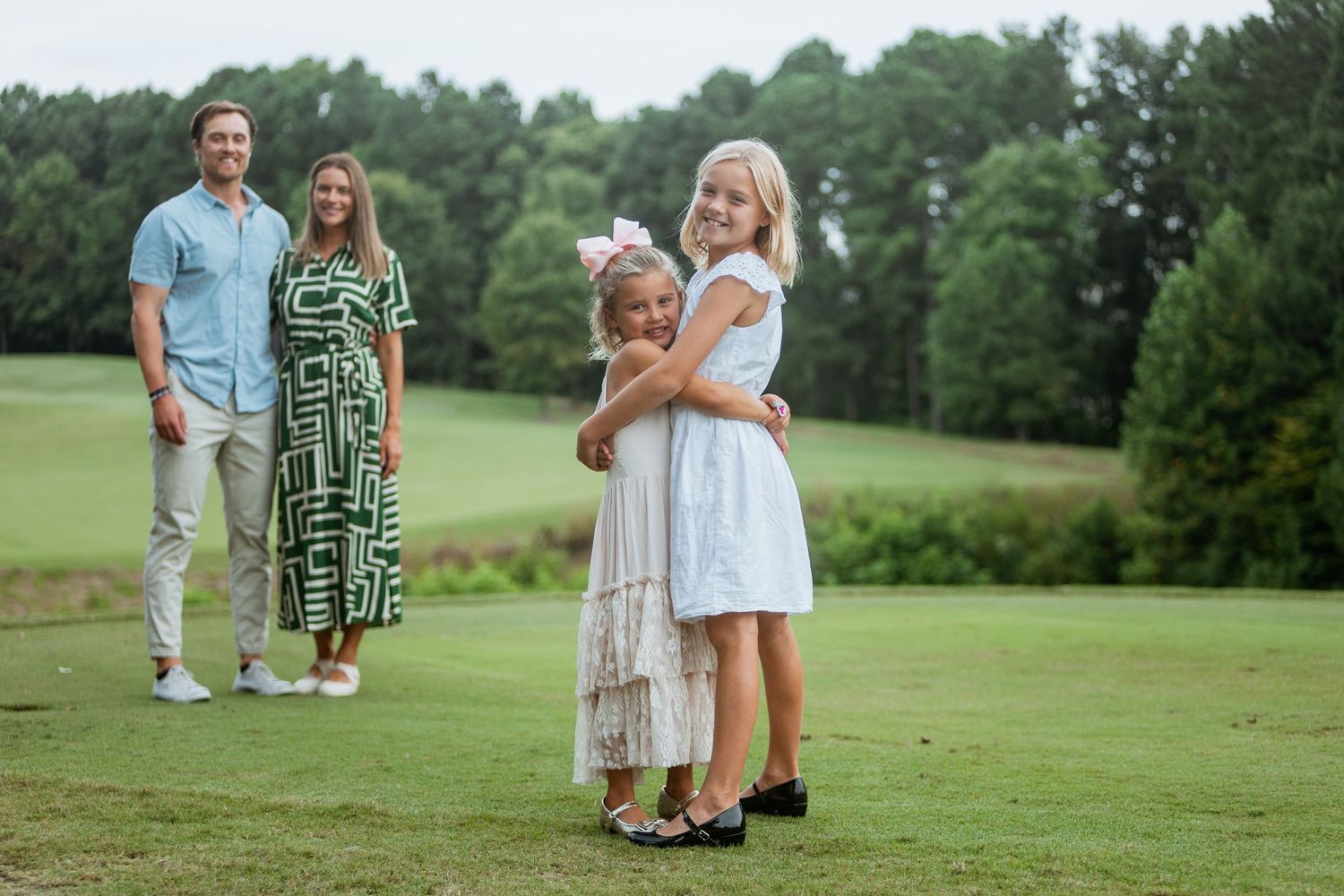 Two girls hugging on a golf course while parents smile in the background.