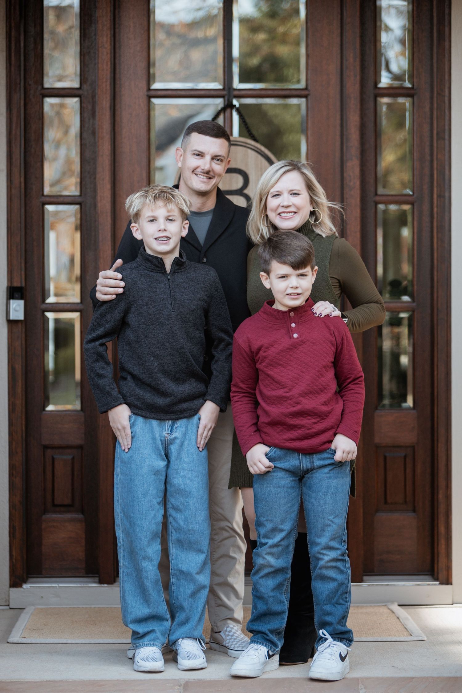 Family of four posing together in front of elegant wooden double doors at home.