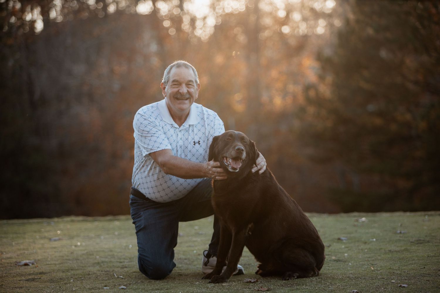 Older man kneeling beside a chocolate Labrador dog for a portrait in a sunlit outdoor setting.