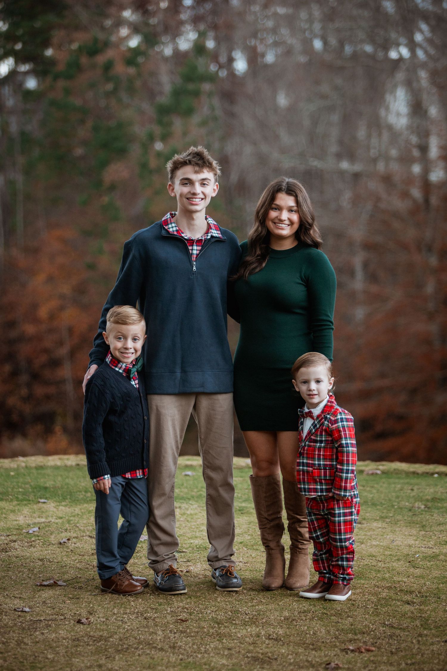 Four siblings in holiday outfits pose on a green lawn with autumn trees in the background.