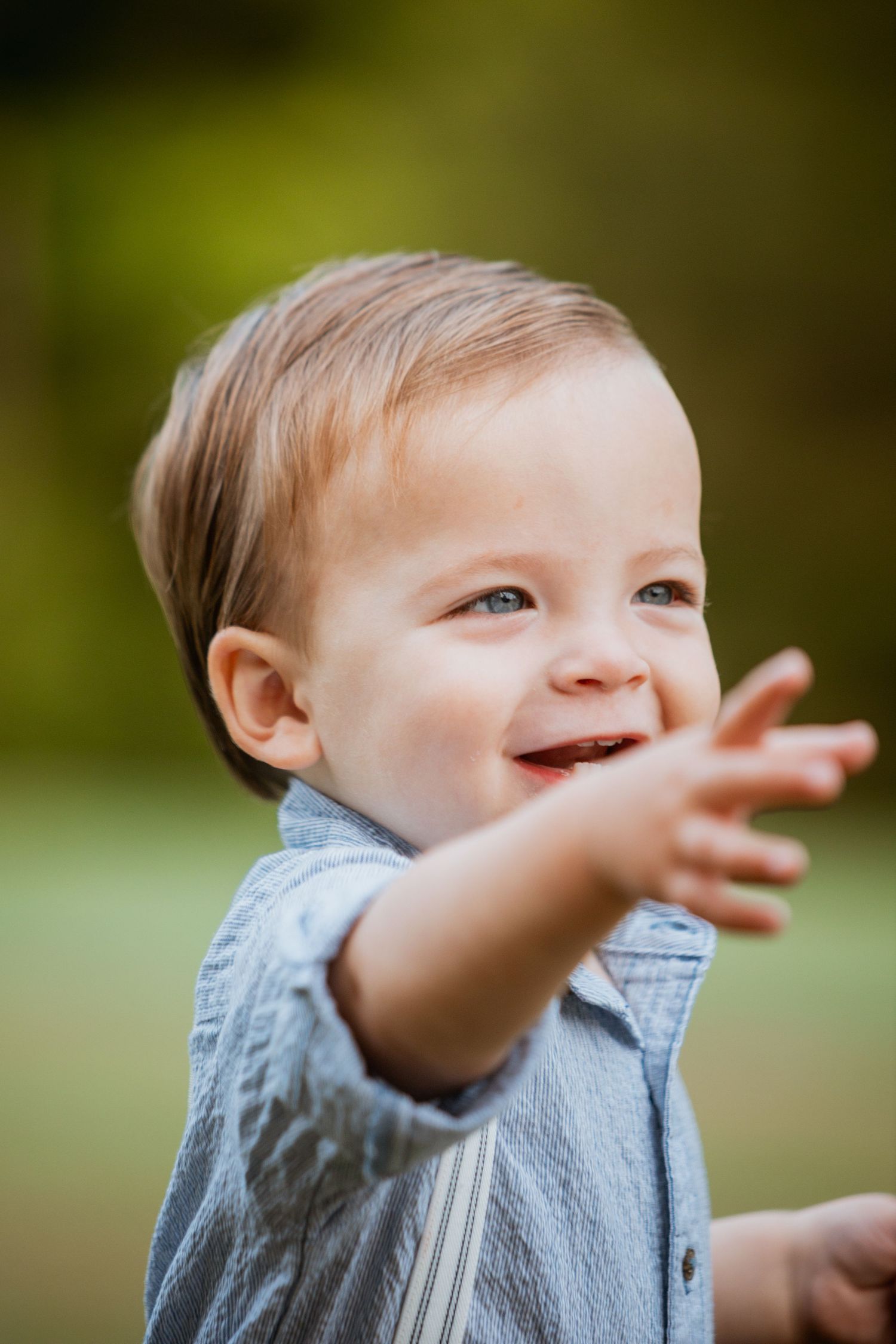 Close-up of a joyful toddler boy in a blue shirt reaching out his hand and smiling outdoors.