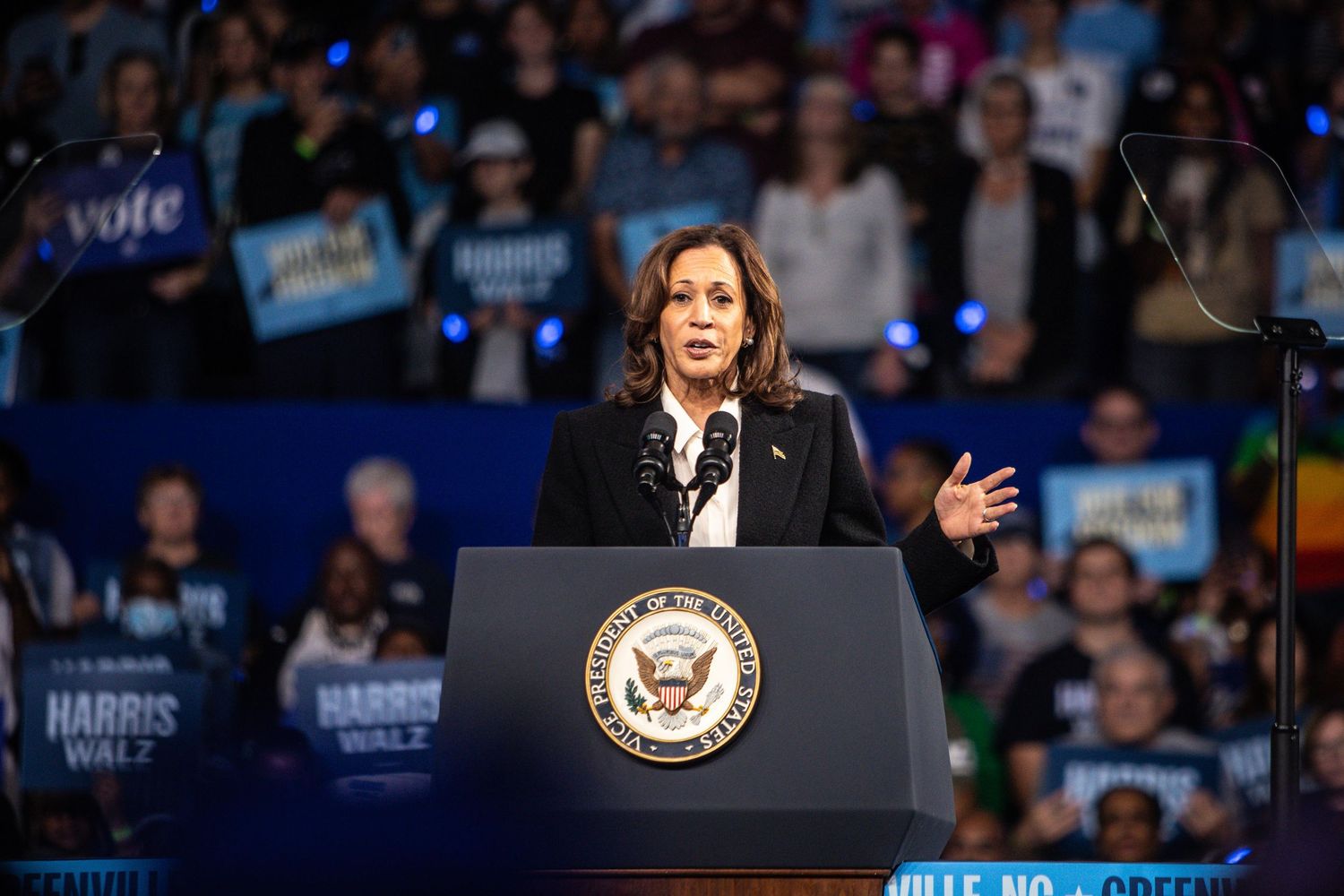 A woman in a black blazer speaks passionately at a Harris-Walz campaign rally podium before a large crowd.